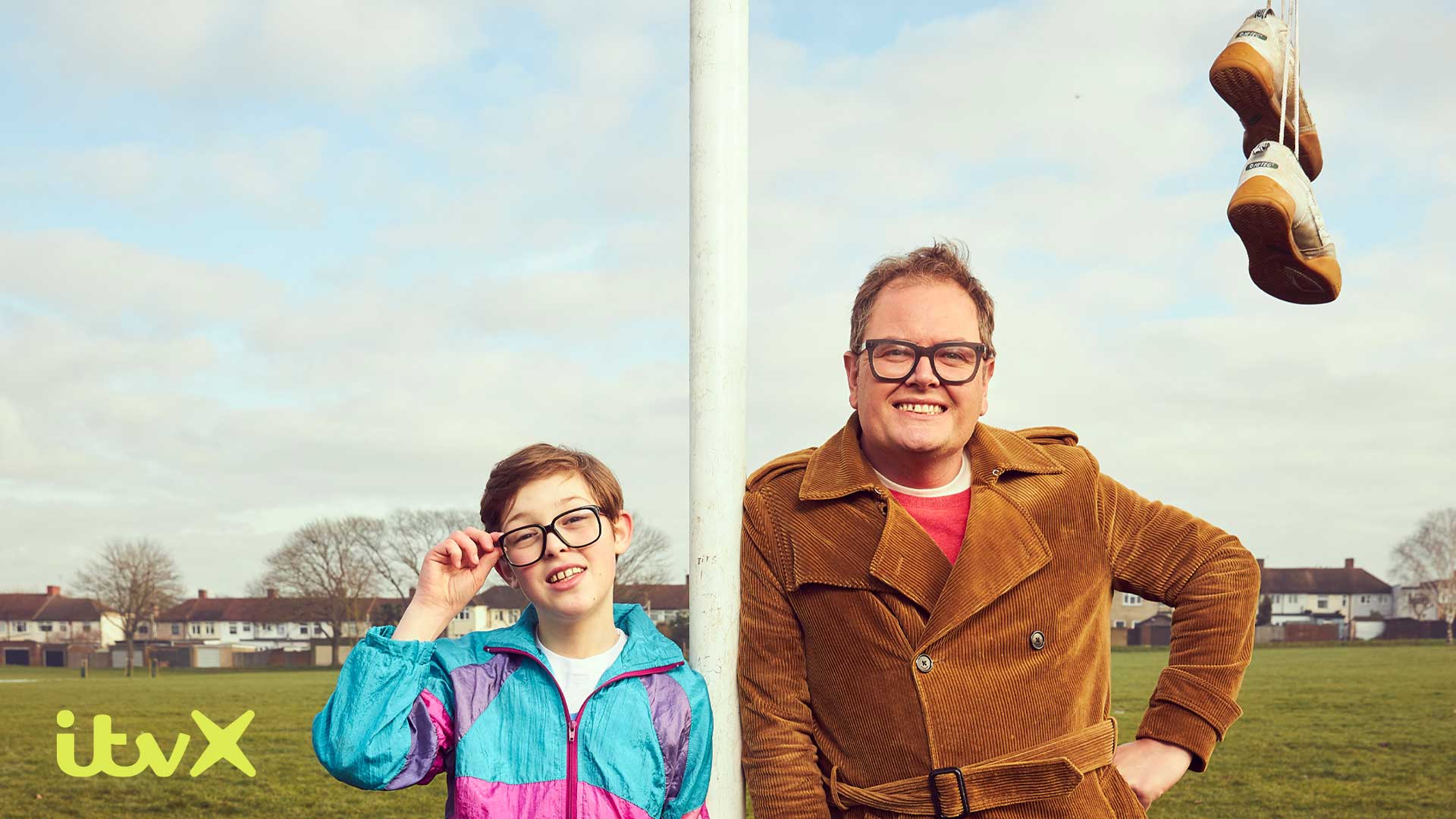 Alan Carr leaning against a goal post in a brown corduroy coat. A young Alan in a shellsuit on his left